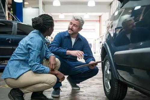 Two mechanics in a garage discuss while inspecting a car. One holds a clipboard, both wearing work attire.