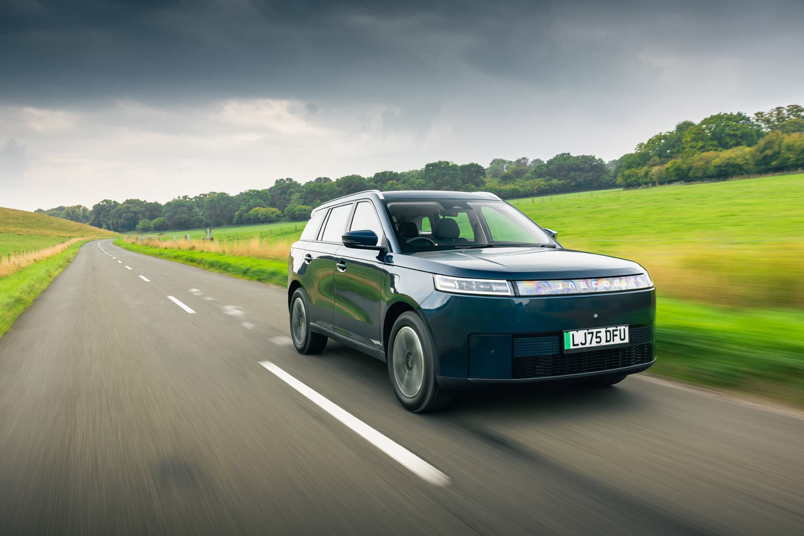 Blue JAECOO E5 driving on a winding country road with green fields and a cloudy sky in the background.