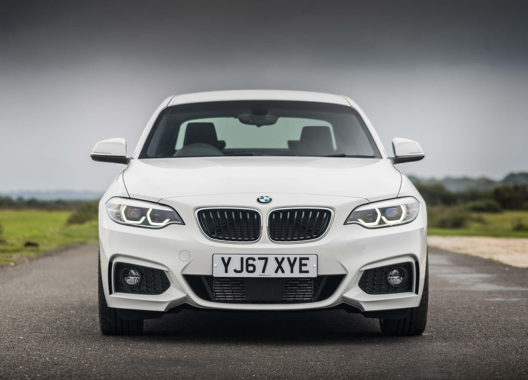 Front view of a white BMW 2 Series on a road, with distinctive headlights and grille, under a cloudy sky.