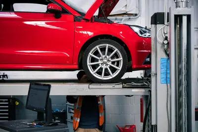 Red car on a lift in a garage, hood open. Mechanic in orange vest partially visible underneath. Computer and tools nearby.