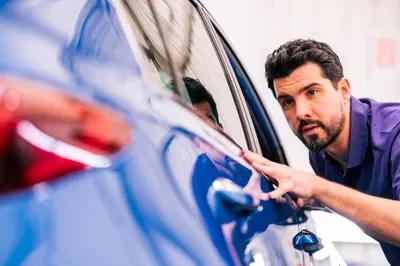 Man inspecting the side of a blue car closely, focusing on the vehicle's surface with a thoughtful expression.