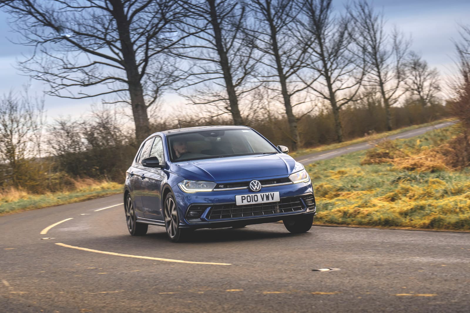 Blue Volkswagen Polo driving on a curved rural road with bare trees and grass in the background under a cloudy sky.
