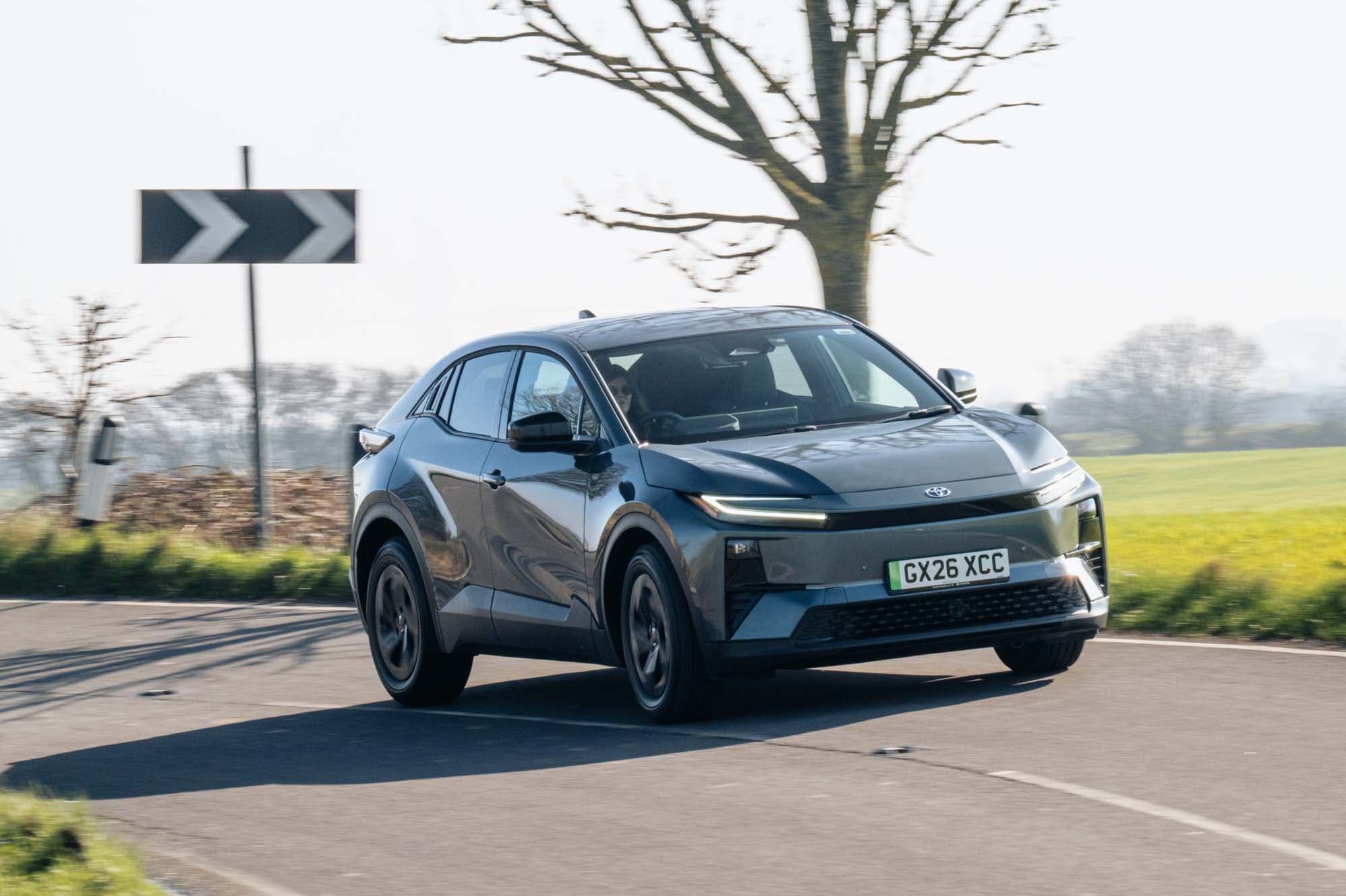 Grey Toyota C-HR+ with modern design driving on a rural road, passing a left curve sign, with trees and fields in the background.