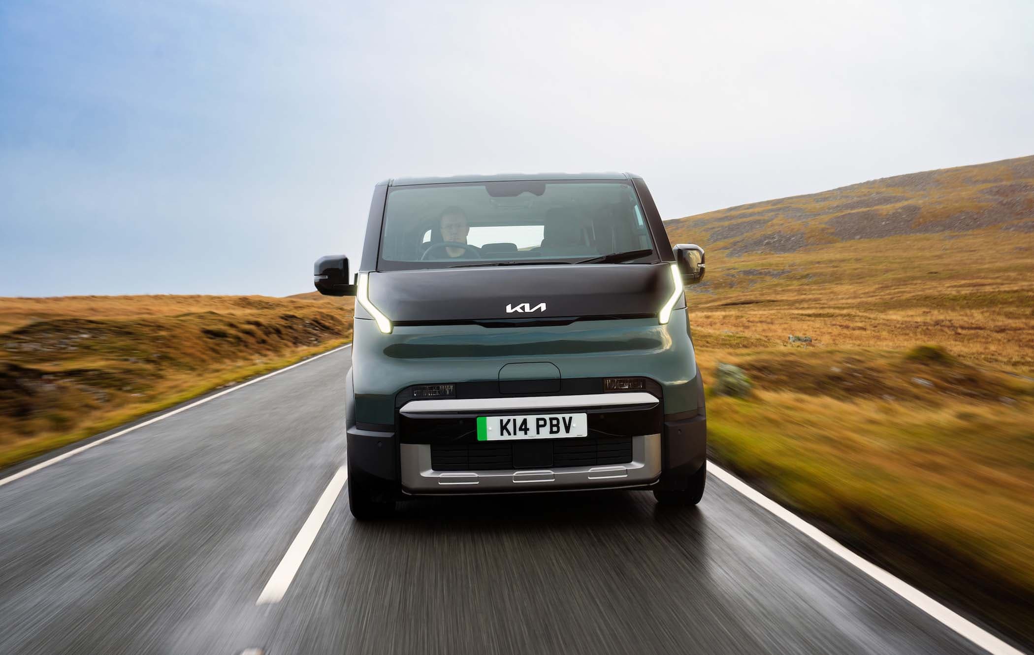 A green Kia PV5 Passenger drives on a scenic, empty road surrounded by grassy hills under a cloudy sky.