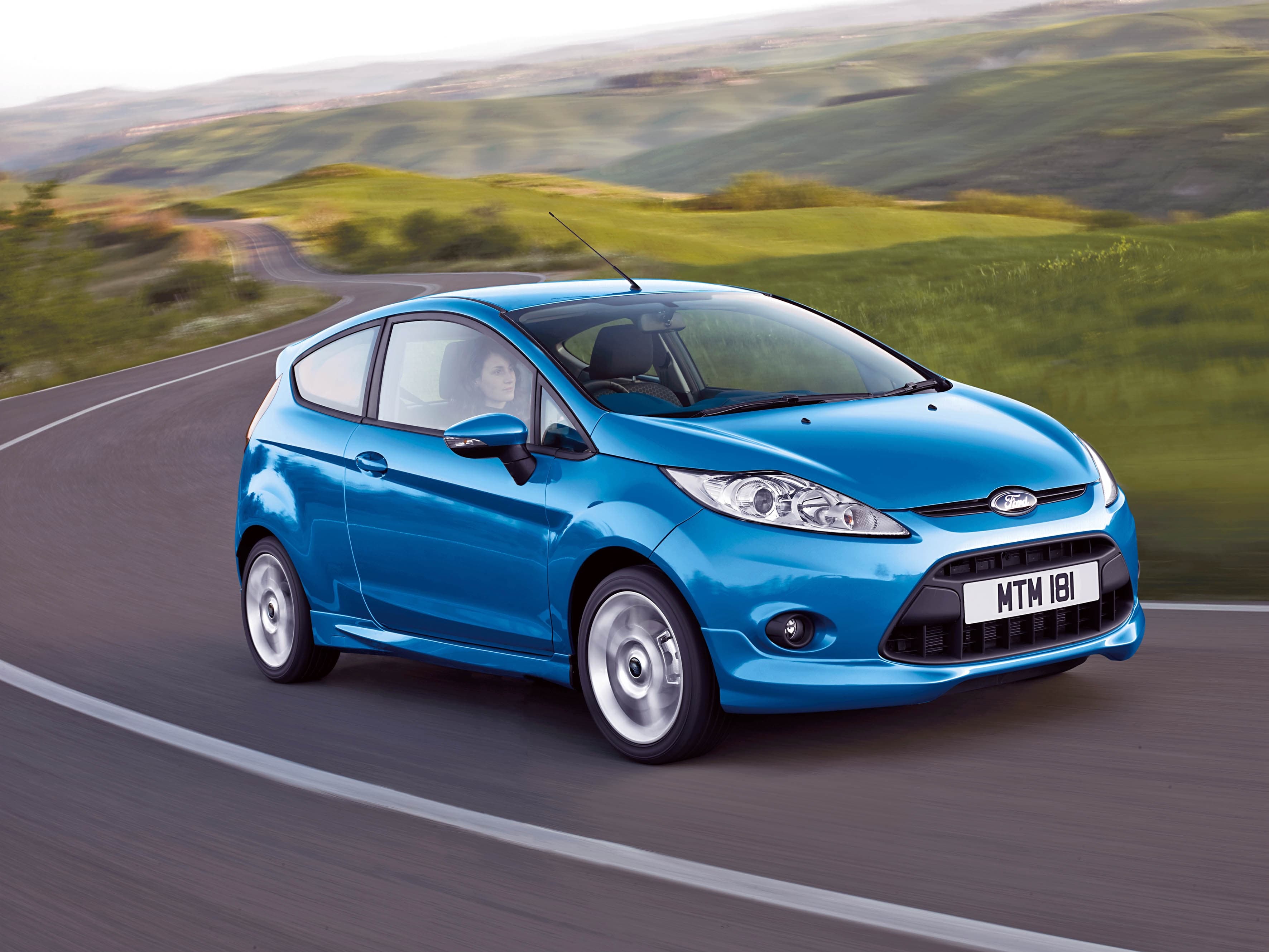 A blue car driving on a winding road through a lush, green hilly landscape under a clear sky.